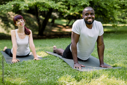 Horizontal shot of young multiethnic couple practicing yoga cobra pose at outdoors summer park. African man and Caucasian girl stretching on yoga practice asana in yoga retreat. Focus on man