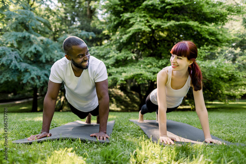 Multiethnic smiling couple, African man and Caucasian girl, doing core workout together in park. Fit young man and woman exercising, doing plank. Sport and fitness outdoors