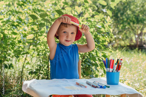 a boy in red Panama sits in nature at a children's table and draws markers on a white watman photo