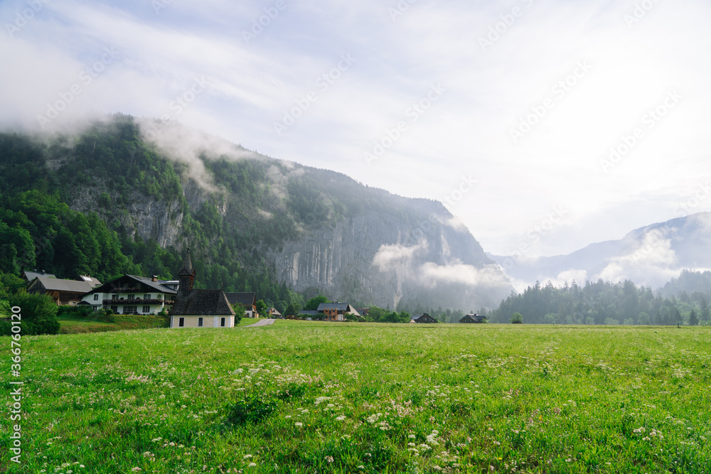 Scenic panoramic view of idyllic church in morning in the alps. Beautiful foggy morning scenery in Alps region, Austria. Great morning view of foggy mountains, fog, house and green meadow in Austria.