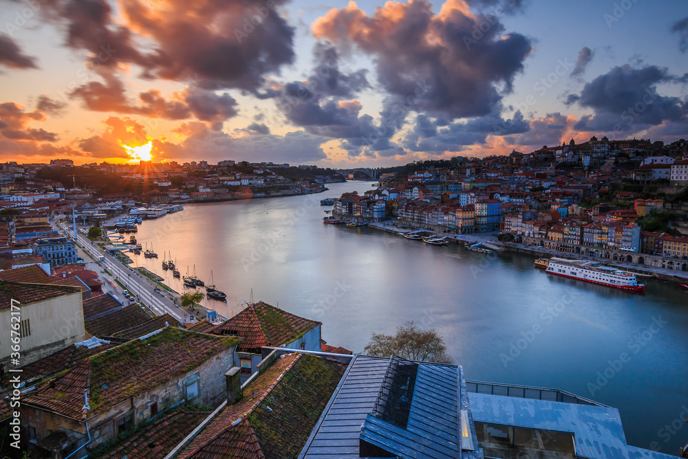Sunset over the rooftops of the city of Porto. River Douro in the Portuguese coastal city. Clouds and sun rays in the evening. City view from an elevated position with harbor, river boats and hills