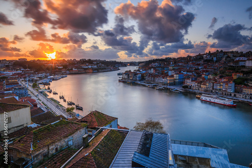 Sunset over the rooftops of the city of Porto. River Douro in the Portuguese coastal city. Clouds and sun rays in the evening. City view from an elevated position with harbor, river boats and hills