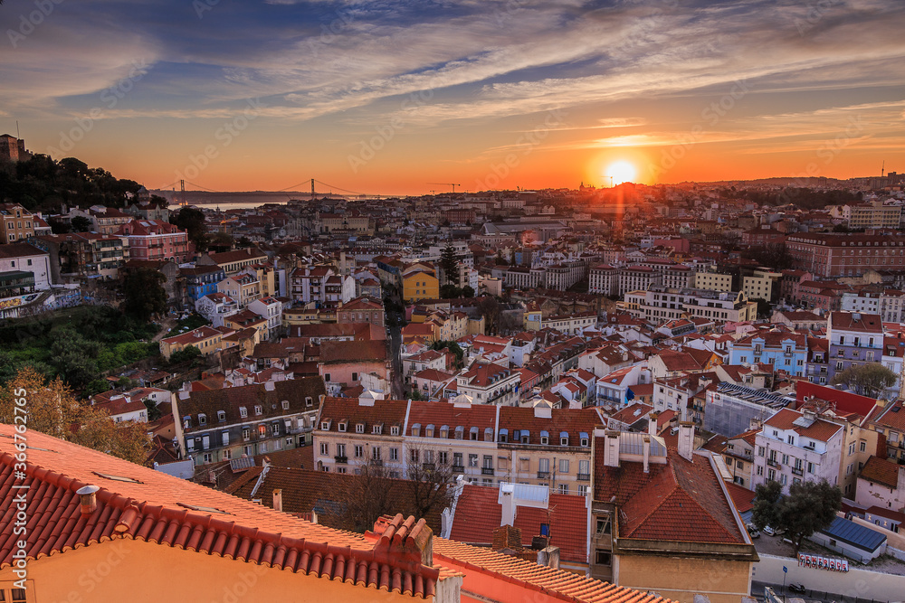 Sunset over the rooftops of the city of Lisbon. Skyline of the old town of the Portuguese capital. Clouds and sun rays in the evening. City view from an elevated position with harbor, river course