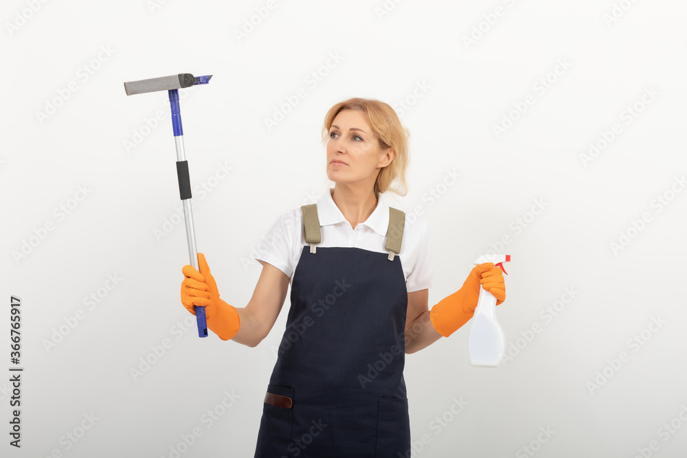 Portrait of cleaning lady or housewife holding a window cleaning brush and a spray. Wearing a uniform apron on light background. European female cleaner ready to work. Portrait of cleaning lady or housewife holding a window cleaning brush and a spray. Wearing a uniform apron on light background. European female cleaner ready to work.