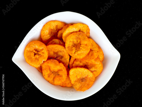 Orange color banana chips with hot red chili flavor in a white bowl against black background
