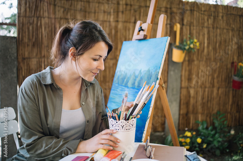 Female painter holding pot with paintbrush in front of painting. Brunette woman holding pot with paint brushes while sitting in outdoor atelier.