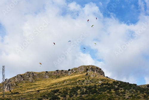 Many paragliders on bright colored parachutes fly together over the mountain - Group of paragliders flying in sunny day against the background of clouds - Adventure, sport, and freedom concept