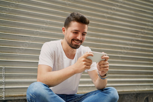 The young man sitting in front of the gray background is playing games with his mobile phone, holding his phone horizontally. The man is smiling while playing games with his mobile phone.   