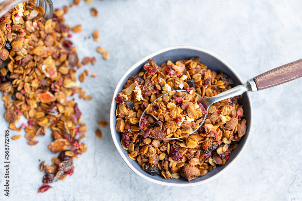 Homemade granola or muesli made of oats, nuts, honey, cinnamon, fruits on white table, blue bowl with spoon. Beautiful healthy snack on breakfast. Top view, flat lay, copy space. Selective focus. Homemade granola or muesli made of oats, nuts, honey, cinnamon, fruits on white table, blue bowl with spoon. Beautiful healthy snack on breakfast. Top view, flat lay, copy space. Selective focus.