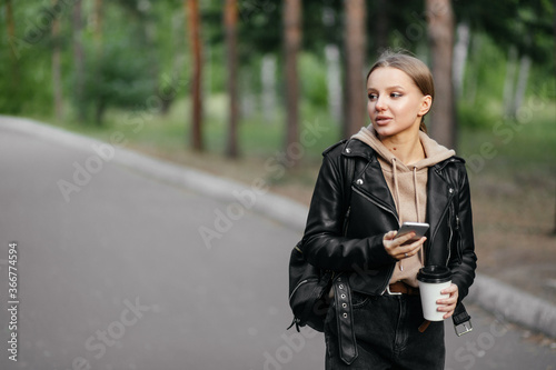 A beautiful woman in a leather jacket walks in the park and smiles pretty while looking to the side with a phone and coffee in her hands. Walking outdoors after a long period of pandemic.