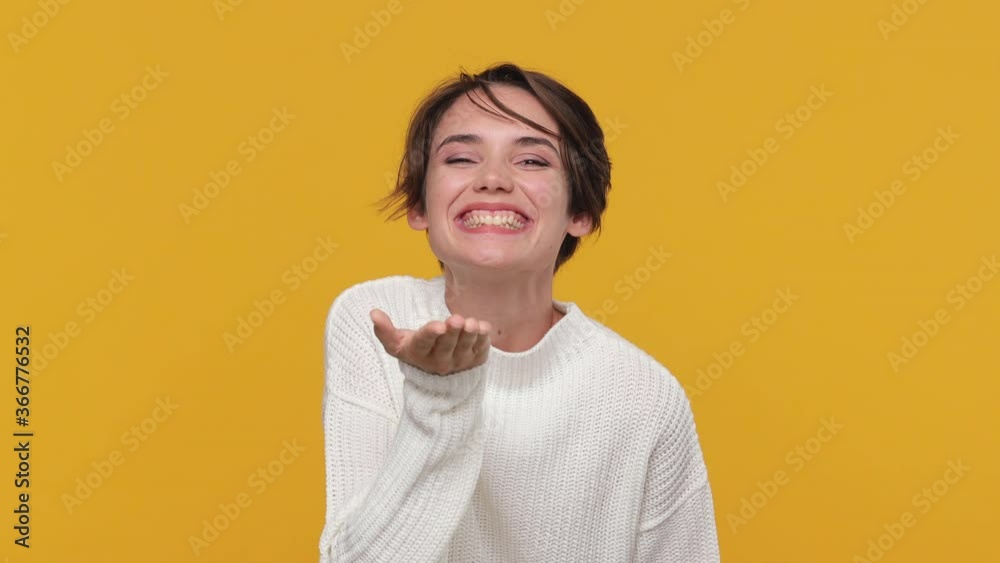Beautiful cute young woman girl 20s years old in white sweater posing isolated on yellow background in studio. People lifestyle concept. Look camera send blow air kiss showing shape heart with hands