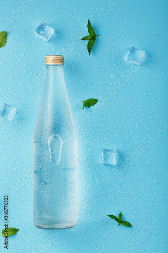 Bottle with an ice cold beverage, ice cubes, drops and mint leaves on a blue background.