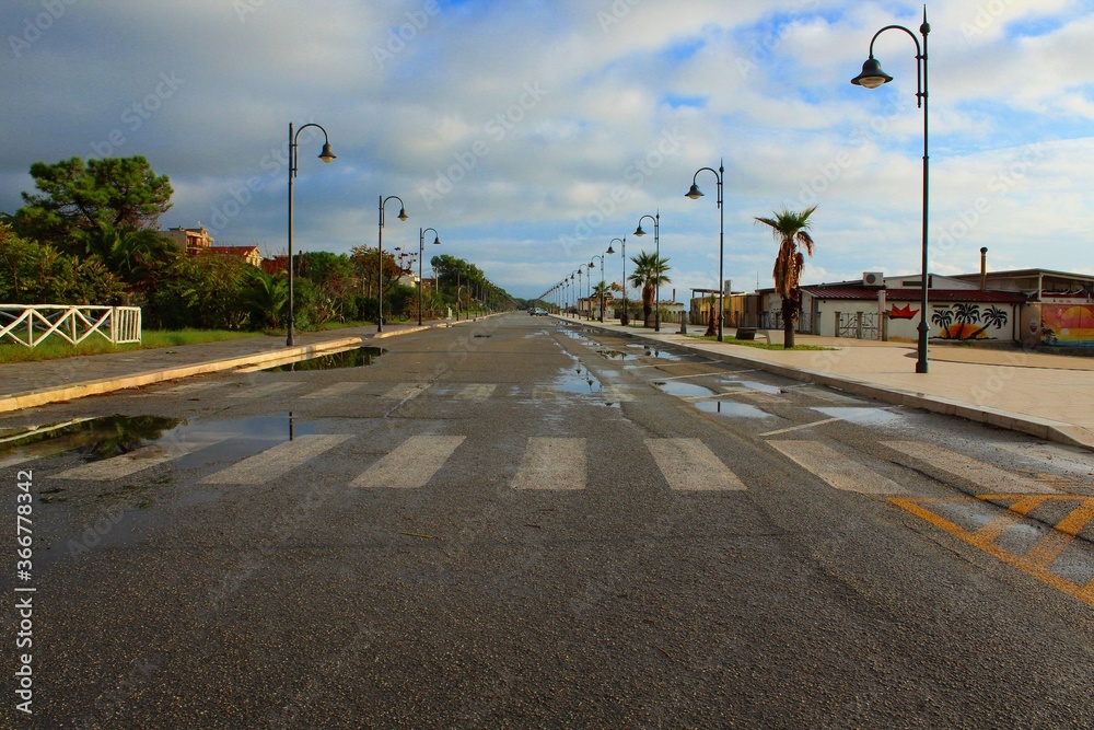 waterfront street is wet after early morning rain, Calabria ( Ardore) , puddles along the empty road and crosswalk, perspective with rows of street lamps along the sidewalks, cloudy blue sky