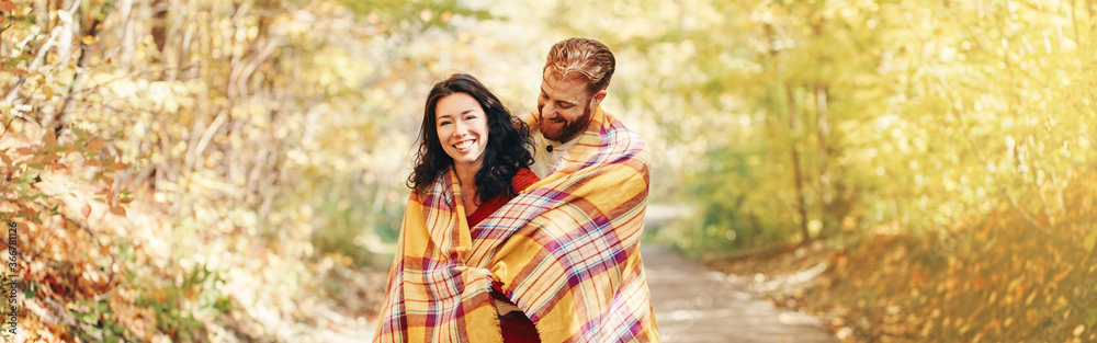 Beautiful couple man woman in love. Boyfriend and girlfriend wrapped in yellow blanket hugging together in park on autumn fall day. Authentic real people. Web banner header for a website.