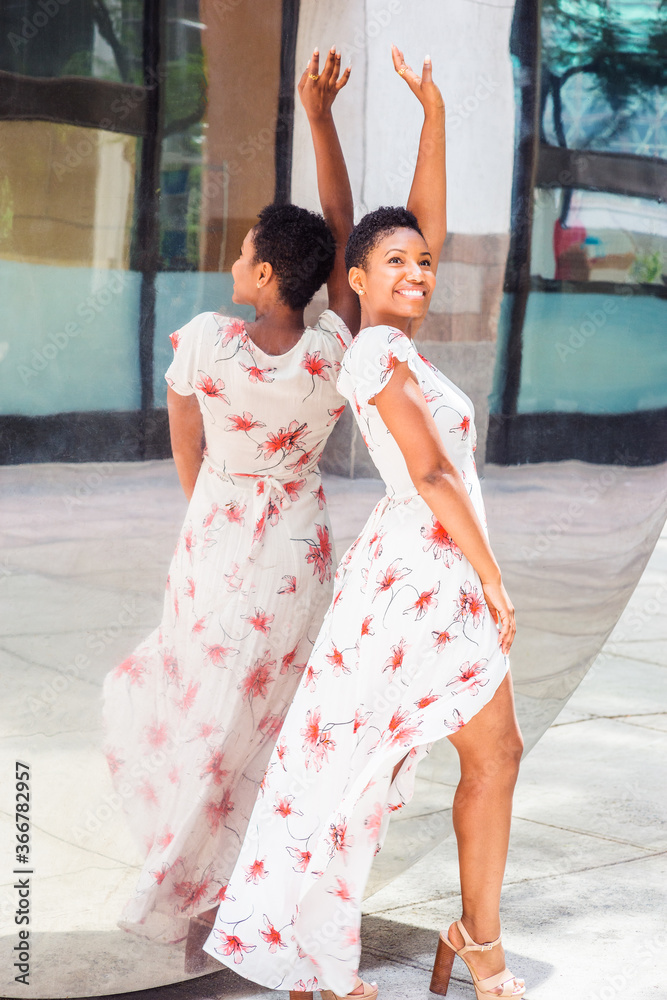 Young Happy African American Woman with short afro hair, wearing long white patterned dress, sandals, standing against mirror outside in New York City, raising arm, big smiling. .