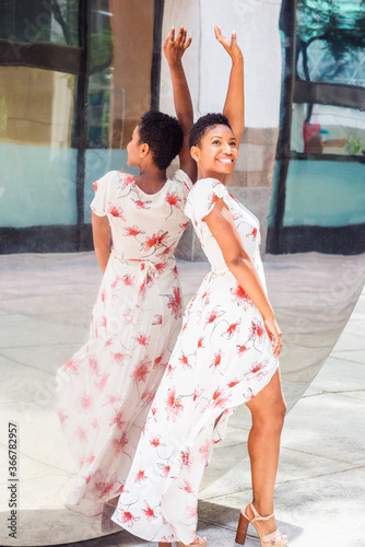 Obraz na płótnie Young Happy African American Woman with short afro hair, wearing long white patterned dress, sandals, standing against mirror outside in New York City, raising arm, big smiling. . w salonie