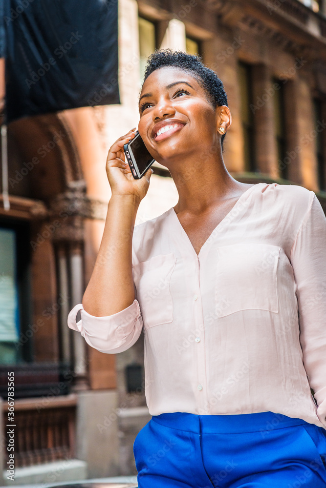 Young Happy African American Woman with short afro hair, traveling in New York City, wearing pink v neck shirt, blue pants, walking on street, talking on cell phone, looking up, smiling..