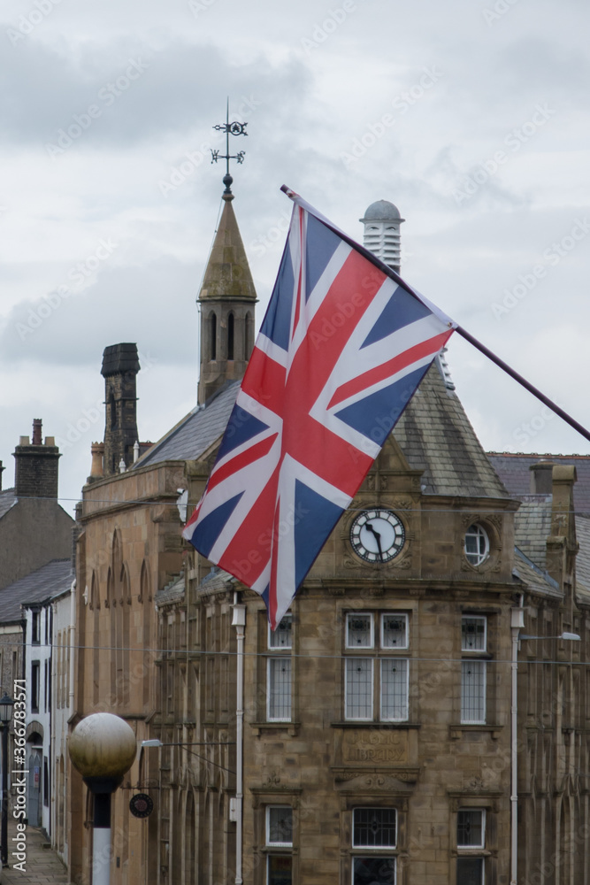 Union Jack flag flying from a pole on the exterior of a building. Old clock tower in the background
