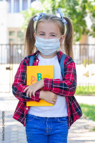 happy schoolgirl in a protective mask with a backpack and a textbook in her hands. in a white T-shirt and a plaid shirt. vertical photo © Elena