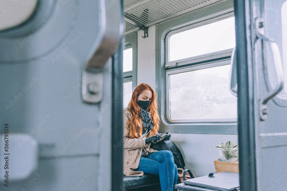 Red-haired girl sits in a train carriage, with a black medical mask, holding a smartphone in her hands. Red-haired girl sits in a train carriage, with a black medical mask, holding a smartphone in her hands.