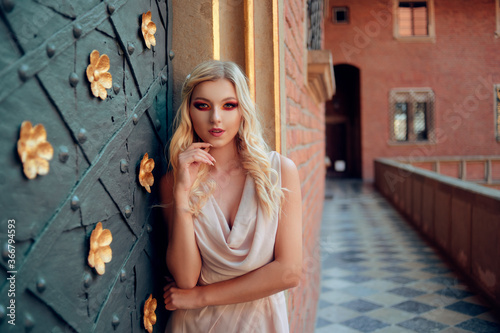 Beautiful pretty blonde with rich professional makeup in the castle. Girl in a dress near the door, golden historical castle. A rich princess in a fairy tale