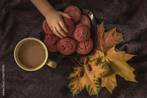 Children's hand reaching for a plate of cranberry cookies. Still life with a cup of coffee, autumn multi-colored maple leaves and acorns on the background of a purple plaid. Autumn concept. Top view