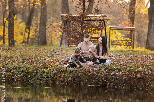 young dad and mom with baby girl and little son having fun on blanket near pond in autumn park on sunny day. happy family concept. mother's, father's, baby's day.
