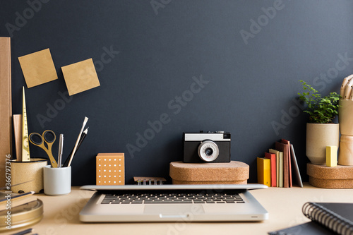 Creative desk with a laptop closed, desk objects, office supplies, books, and plant on a dark blue background.	