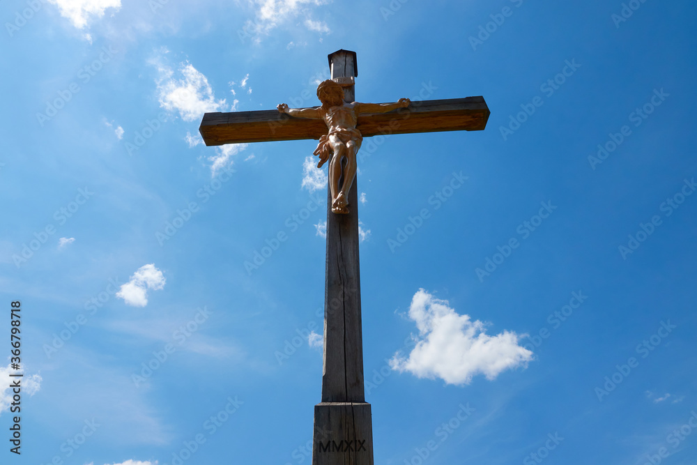 Jesus figure ( cruci fixus ) on brown wooden cross, Roman numerals on the wood. Blue sky with white clouds. Germany.