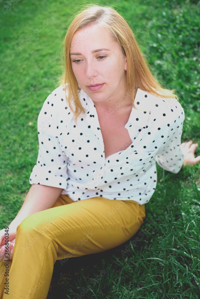 beautiful middle age woman sitting on a grass. thoughtful girl wearing trendy polka dot blouse and yellow pants. calm and pensive woman. beautiful middle age woman sitting on a grass. thoughtful girl wearing trendy polka dot blouse and yellow pants. calm and pensive woman.