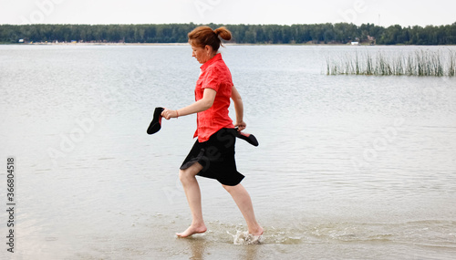 A young businesswoman in office clothes runs barefoot on the water on a lake in the heat and holds high-heeled shoes in her hands.