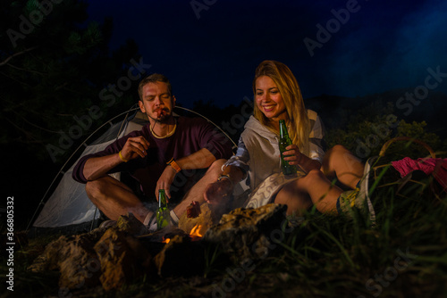 Young couple baking sausages on the campfire and drinking beer in the forest hill in the dusk