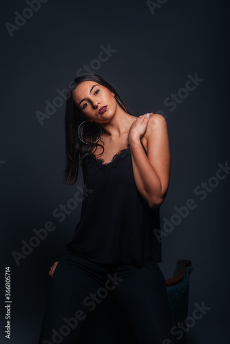 Sexy brunette wearing a black tank top and black pants posing while sitting on the top edge of a chair inside a studio on a dark background