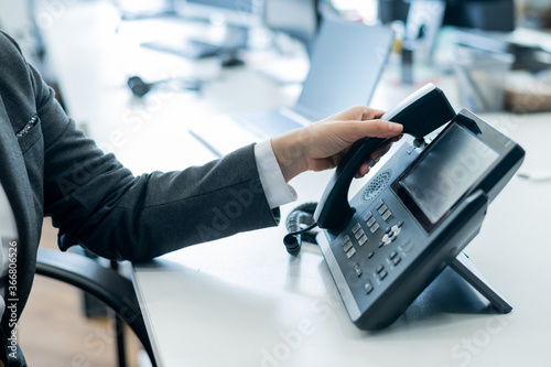 Closeup female hand on landline phone in office. Faceless woman in a suit works as a receptionist answering the phone to customer calls. photo