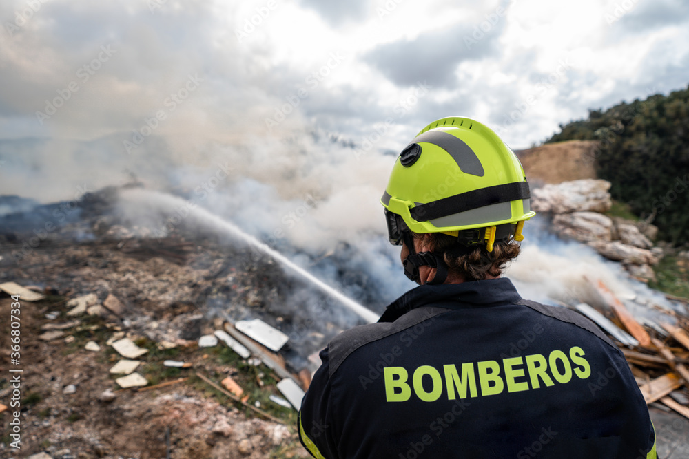 Back view of brave fireman in protective uniform standing with hose and extinguishing fire on dump in mountains Back view of brave fireman in protective uniform standing with hose and extinguishing fire on dump in mountains