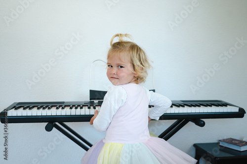 Back view of schoolgirl in fluffy skirt sitting at synthesizer and preparing for music class looking at camera photo