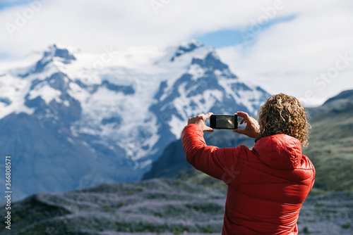 Back view of unrecognizable male tourist in warm clothes taking pictures of amazing mountain ridge while using cellphone camera and travelling around Iceland during vacation in winter photo