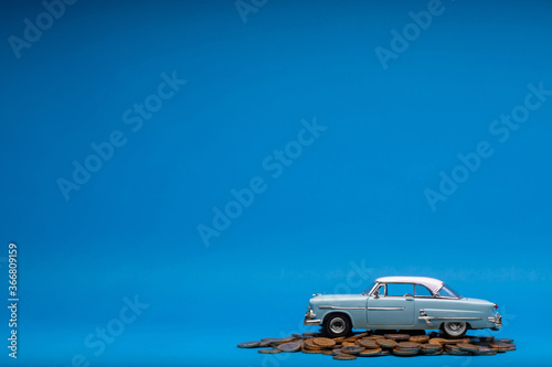 Light blue car figurine placed on top of a large heap of coins, on blue background.