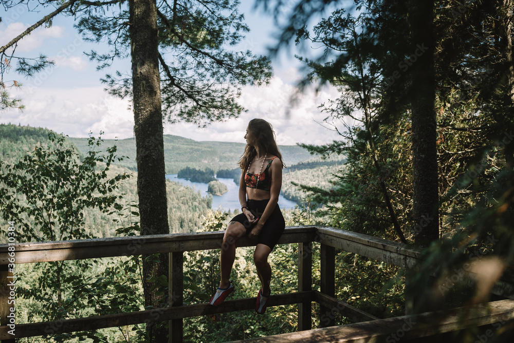 Full body of young female tourist sitting on railing of wooden terrace and looking away in forest of La Mauricie National Park in Quebec, Canada Full body of young female tourist sitting on railing of wooden terrace and looking away in forest of La Mauricie National Park in Quebec, Canada