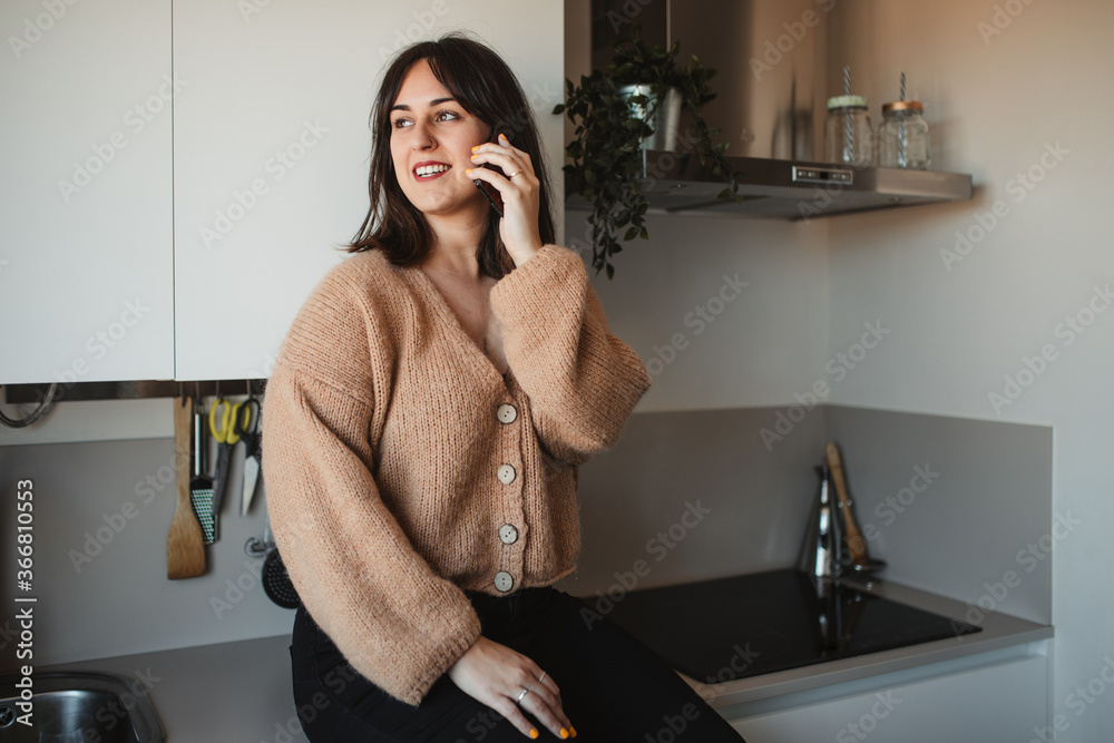 Delighted female in trendy sweater sitting in kitchen countertop and having a conversation on a mobile phone Delighted female in trendy sweater sitting in kitchen countertop and having a conversation on a mobile phone