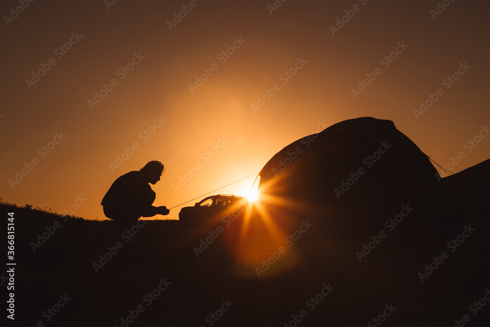 A man is silhouetted against the orange sky of sunset or dawn. A man sets up a camp in the mountains near the car. Journey. A man is silhouetted against the orange sky of sunset or dawn. A man sets up a camp in the mountains near the car. Journey.