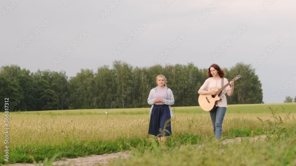 two young beautiful girls walk and sing along a wheat field at sunset on a summer evening. The girl accompanies the guitar two young beautiful girls walk and sing along a wheat field at sunset on a summer evening. The girl accompanies the guitar