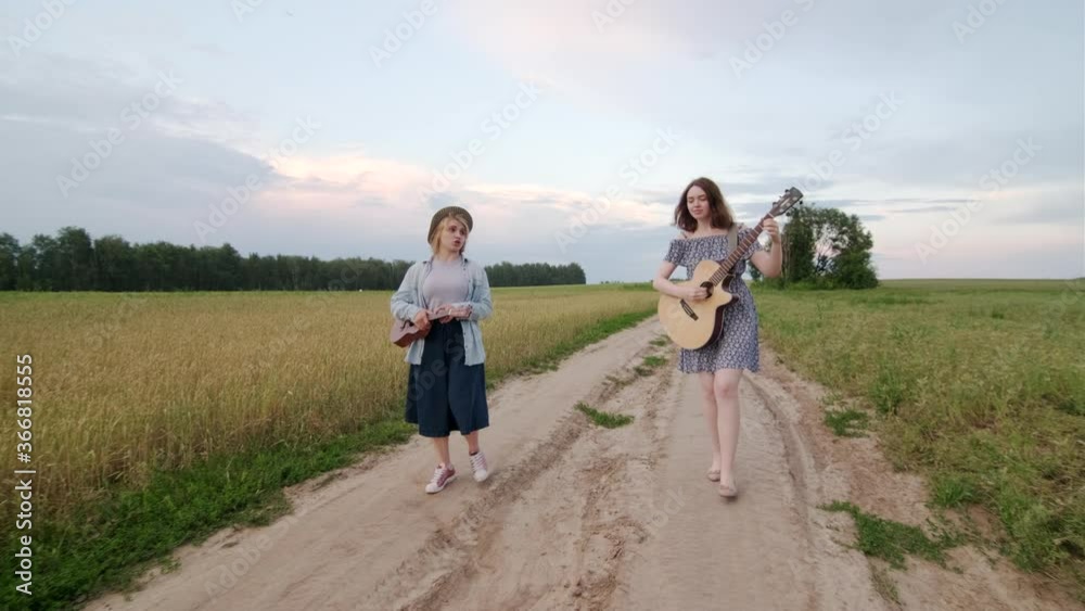 duet of cute cheerful girls. Wheat field road at sunset in summer evening. Guitar and ukulele. Girls sing and laugh. Dynamic camera movement duet of cute cheerful girls. Wheat field road at sunset in summer evening. Guitar and ukulele. Girls sing and laugh. Dynamic camera movement