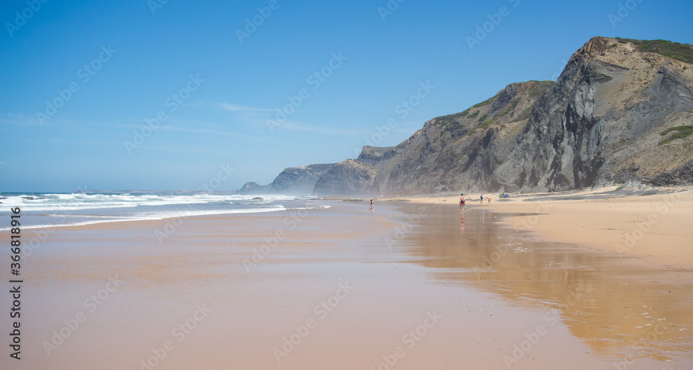 Scenic beach with rare people, large band of golden sand, dark cliff and blue sky. Algarve, Portugal.