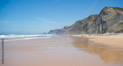 Scenic beach with rare people, large band of golden sand, dark cliff and blue sky. Algarve, Portugal.