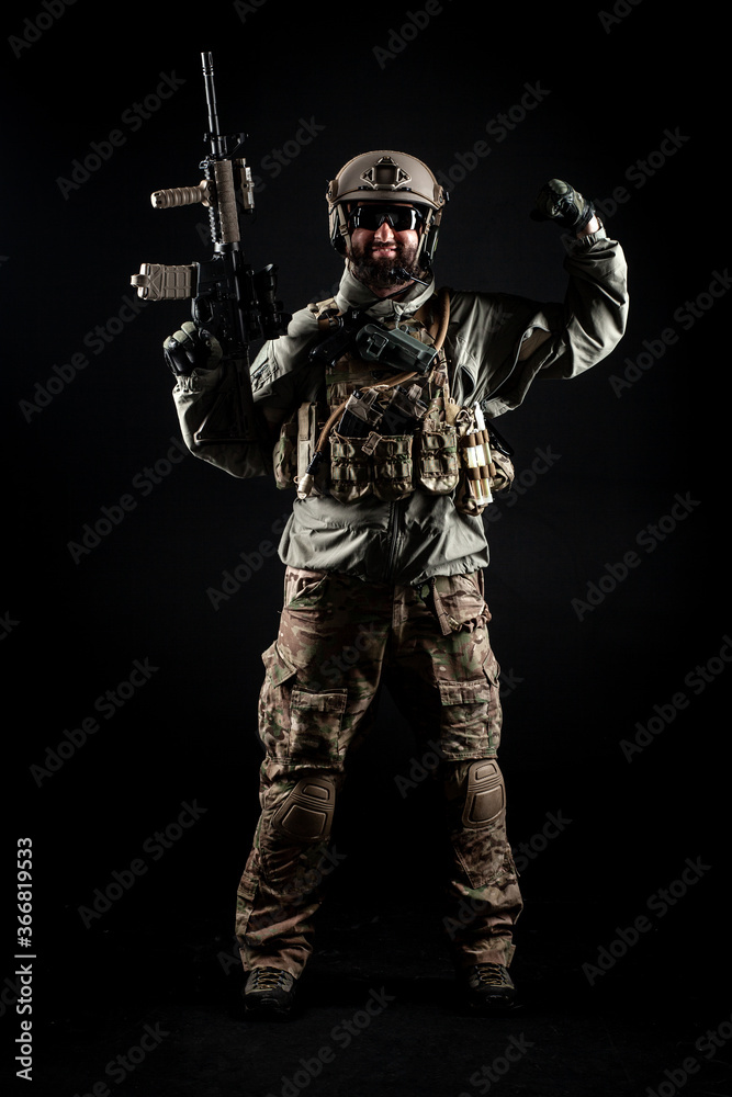 USA soldier in a military suit with a rifle smiles and shows strength against a dark background, American commando in uniform with a gun USA soldier in a military suit with a rifle smiles and shows strength against a dark background, American commando in uniform with a gun