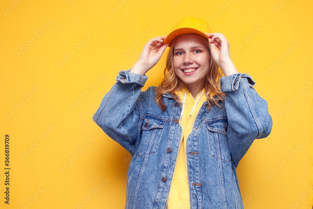 girl in a denim jacket puts on a cap on a yellow background and smiles