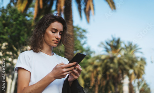 pretty woman writes message on mobile smartphone while walking Park barcelona on a warm summer day, hipser girl listens to music on earphones and searches for information using cell phone photo