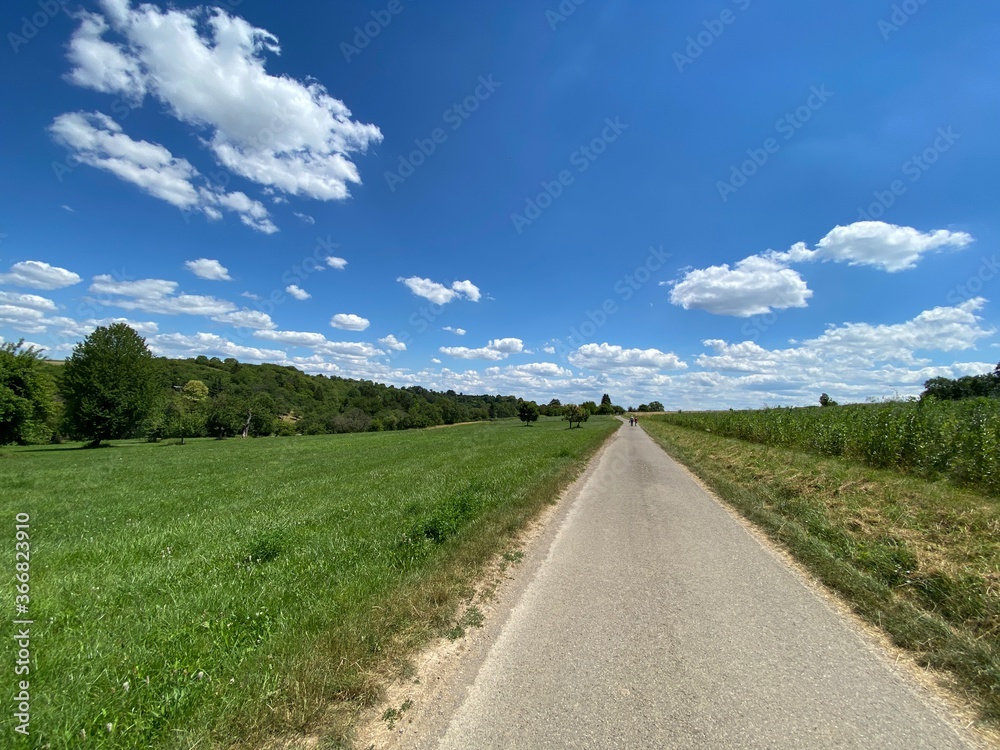 Schmale Straße am Horizont Spaziergänger, daneben grünes Feld. Himmel ist blau mit weißen Wolken.