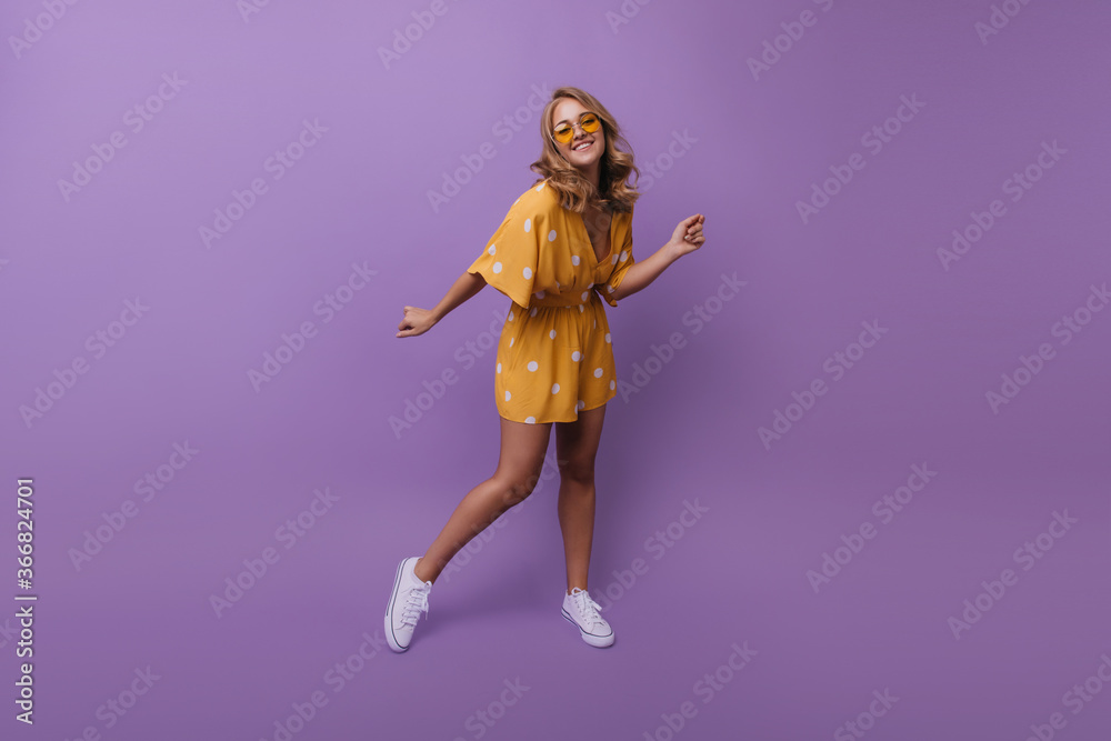 Full-length portrait of happy tanned girl in white sneakers. Studio photo of pleased blonde woman dancing during photoshoot on purple background. Full-length portrait of happy tanned girl in white sneakers. Studio photo of pleased blonde woman dancing during photoshoot on purple background.